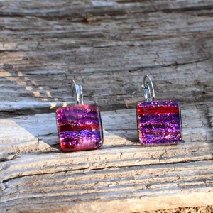 May include: A pair of square dangle earrings with a dichroic glass design. The earrings feature a vibrant mix of red, purple, and iridescent colors. The earrings are suspended from silver-colored lever-back ear wires. The earrings are displayed on a weathered wooden surface.