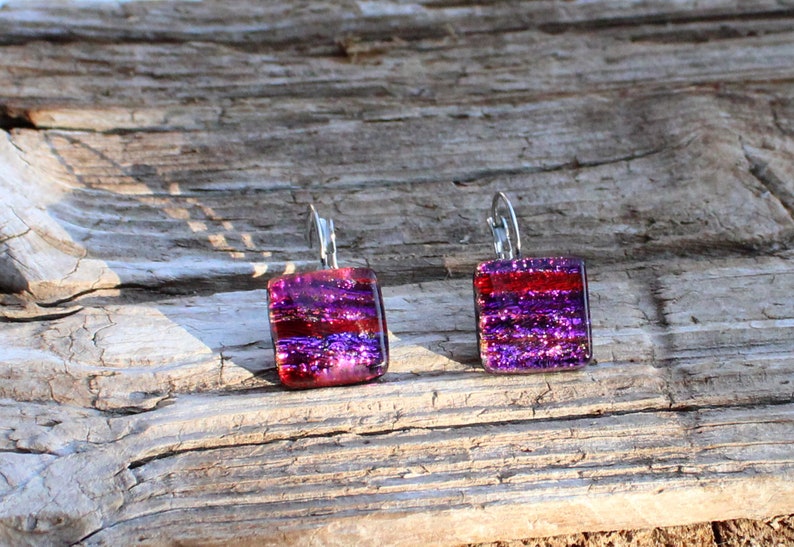 May include: A pair of square dangle earrings with a dichroic glass design. The earrings feature a vibrant mix of red, purple, and iridescent colors. The earrings are suspended from silver-colored lever-back ear wires. The earrings are displayed on a weathered wooden surface.
