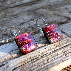 May include: A pair of square dangle earrings with a dichroic glass design. The earrings feature a vibrant mix of red, pink, and purple hues with a metallic shimmer. The earrings are set on silver-colored lever-back ear wires.
