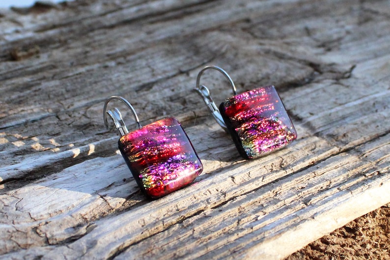 May include: A pair of square dangle earrings with a dichroic glass design. The earrings feature a vibrant mix of red, pink, and purple hues with a metallic shimmer. The earrings are set on silver-colored lever-back ear wires.
