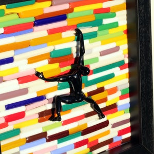 K&ouml;nnte beinhalten: Eine schwarze Figuren-Skulptur klettert an einer Wand aus bunten Glasfliesen. Die Wand besteht aus vielen verschiedenen Farben, darunter Rot, Gelb, Gr&uuml;n, Blau und Lila. Die Figur befindet sich in einer Kletterpose, mit einem Arm nach oben und dem anderen Arm nach unten gestreckt. Die Figur befindet sich in einem schwarzen Rahmen.