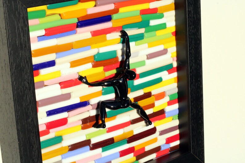 K&ouml;nnte beinhalten: Eine schwarze Figuren-Skulptur klettert an einer Wand aus bunten Glasfliesen. Die Wand besteht aus vielen verschiedenen Farben, darunter Rot, Gelb, Gr&uuml;n, Blau und Lila. Die Figur befindet sich in einer Kletterpose, mit einem Arm nach oben und dem anderen Arm nach unten gestreckt. Die Figur befindet sich in einem schwarzen Rahmen.
