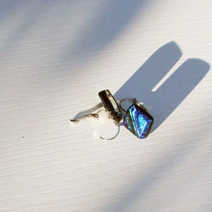 May include: A pair of silver-toned dangle earrings with a dichroic glass design. The earrings feature a square-shaped, iridescent blue and green glass pendant. The earrings are displayed on a white surface with a shadow.