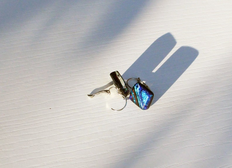 May include: A pair of silver-toned dangle earrings with a dichroic glass design. The earrings feature a square-shaped, iridescent blue and green glass pendant. The earrings are displayed on a white surface with a shadow.