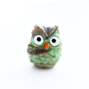 May include: A small green glass owl figurine with brown and white accents. The owl has large white eyes with black pupils and an orange beak.