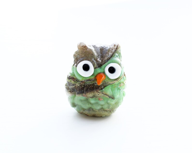 May include: A small green glass owl figurine with brown and white accents. The owl has large white eyes with black pupils and an orange beak.