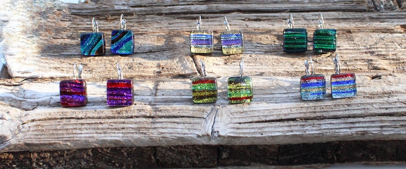 May include: A collection of square dichroic glass earrings with silver-colored ear wires. The earrings feature various colors, including blue, green, red, purple, and gold, with striped and textured patterns. The earrings are displayed on a weathered wooden surface.
