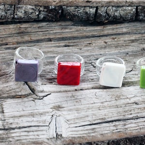 May include: Five glass rings with square glass inlays. The inlays are orange, purple, red, white, and green.