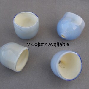 May include: Four small, light blue glass cups with a white interior and a blue rim. The cups are arranged in a semi-circle on a gray surface. The text "7 colors available" is written in black below the cups.