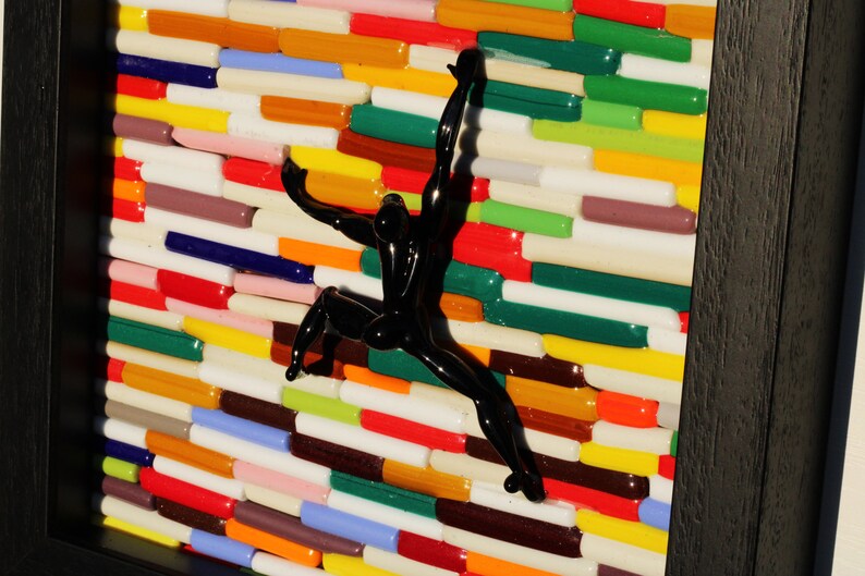 K&ouml;nnte beinhalten: Eine schwarze Glasskulptur einer Person, die eine Wand aus bunten Glasfliesen erklimmt.