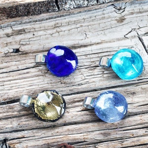 Könnte beinhalten: Vier Glasanhänger mit silbernen Metallscharnieren. Die Anhänger sind rund und haben verschiedene Farben und Muster: Blau, Türkis, Gold und Hellblau.