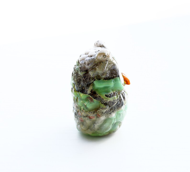 May include: A green, brown, and white owl figurine with a small orange beak. The owl is made of glass and has a textured surface.