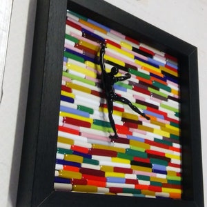 K&ouml;nnte beinhalten: Ein schwarzes gerahmtes Kunstwerk mit einer schwarzen Silhouette einer Person, die eine Wand aus bunten Glasfliesen erklimmt.