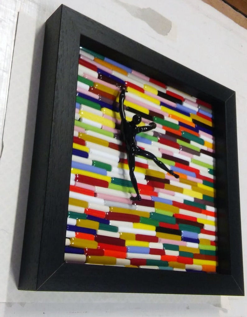 K&ouml;nnte beinhalten: Ein schwarzes gerahmtes Kunstwerk mit einer schwarzen Silhouette einer Person, die eine Wand aus bunten Glasfliesen erklimmt.