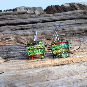 May include: Square dichroic glass earrings with lever-back closures. The earrings feature layers of green, yellow, and red colors. The glass has a shimmering, iridescent quality. The earrings are displayed on a weathered wooden surface.
