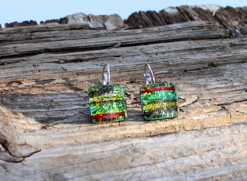 May include: Square dichroic glass earrings with lever-back closures. The earrings feature layers of green, yellow, and red colors. The glass has a shimmering, iridescent quality. The earrings are displayed on a weathered wooden surface.