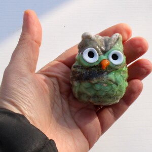 May include: A small green glass owl figurine with white eyes and an orange beak. The owl is sitting in a hand.