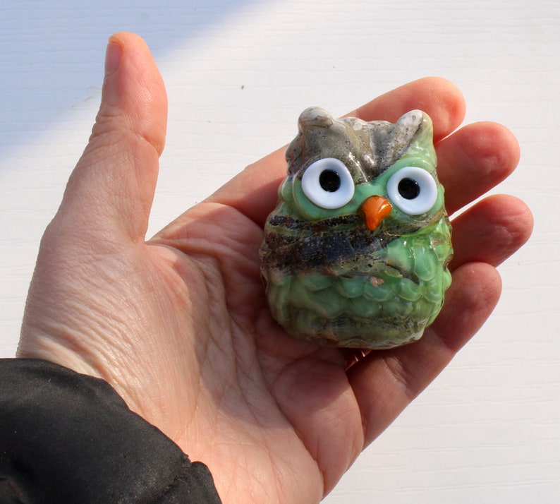 May include: A small green glass owl figurine with white eyes and an orange beak. The owl is sitting in a hand.