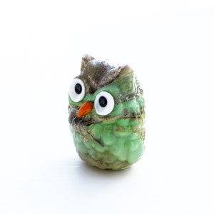 May include: A small green glass owl figurine with large white eyes and an orange beak. The owl is sitting on a white surface.