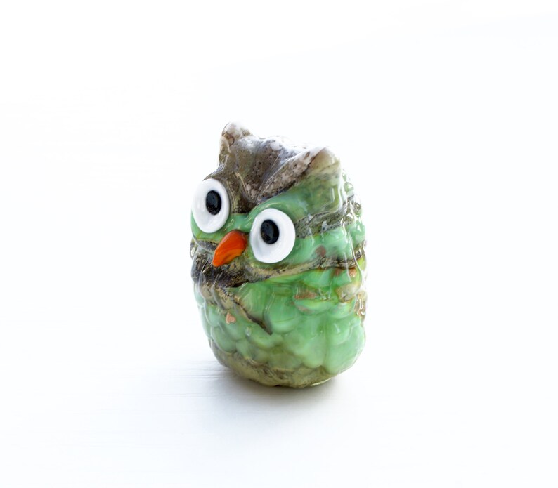May include: A small green glass owl figurine with large white eyes and an orange beak. The owl is sitting on a white surface.