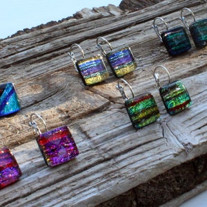 May include: A collection of square dichroic glass earrings in various colors, including blue, green, purple, and red, with silver-tone lever-back closures. The earrings are displayed on a weathered wooden surface.