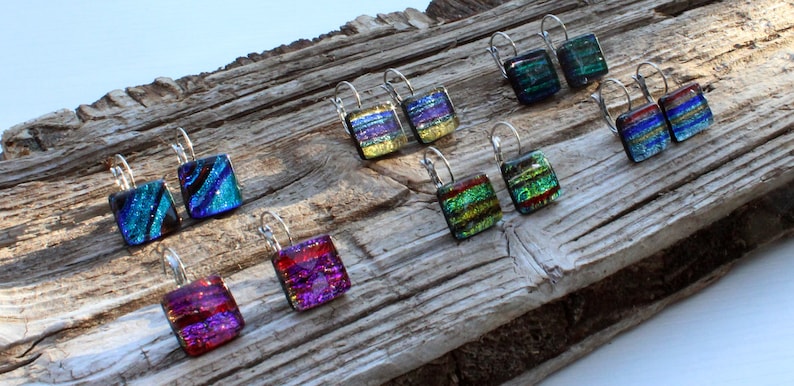 May include: A collection of square dichroic glass earrings in various colors, including blue, green, purple, and red, with silver-tone lever-back closures. The earrings are displayed on a weathered wooden surface.