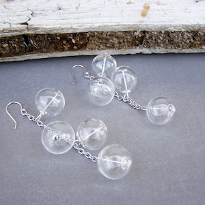 Könnte beinhalten: Ohrringe mit klaren Glasblasen mit silberfarbener Kette und Ohrhaken. Jeder Ohrring hat mehrere klare Glaskugeln unterschiedlicher Größe, die durch eine filigrane Kette verbunden sind. Die Ohrringe liegen auf grauem Untergrund.