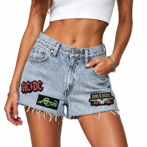 May include: Light blue acid-wash denim shorts featuring a frayed hem and three embroidered patches. The patches include a red AC/DC logo, a green Poison logo, and a black Guns N' Roses logo with a graphic of two pistols and red roses.