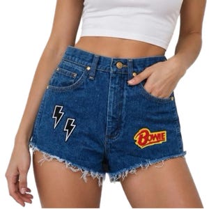 May include: High-waisted blue denim shorts featuring a frayed hemline, a metal button closure, and two embroidered patches. One patch displays two white lightning bolts with black outlines, while the other patch features the red and yellow text Bowie.