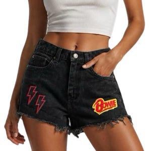 May include: High-waisted black denim shorts featuring a frayed hem, a button and zipper fly, and embroidered patches. The patches include two red lightning bolts on the left side and a yellow and red patch with the text Bowie on the right.