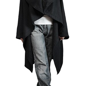 May include: A black, draped coat with a wide collar. The coat is worn over a white shirt and blue jeans. The coat has long sleeves and a flowing design. The person is wearing black shoes.