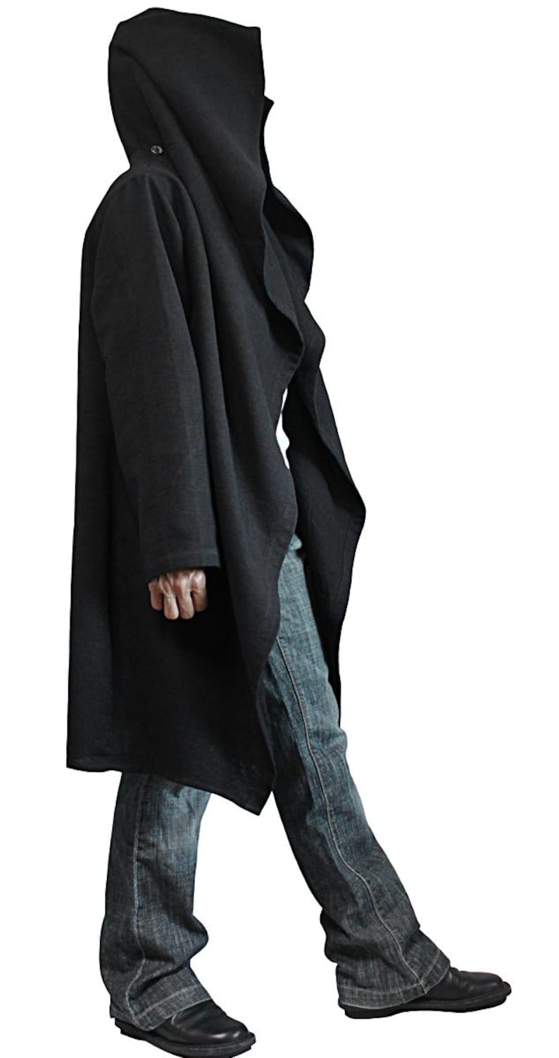 May include: A long, black hooded coat with a draped design. The coat features a large hood and a button closure. The person is wearing blue jeans and black shoes. The coat is made of a soft, flowing fabric.