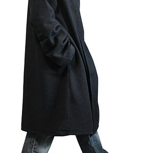 May include: A dark gray hooded coat with a high collar and long sleeves. The coat is knee-length and worn with blue jeans and black shoes. The coat has a relaxed fit and a minimalist design, suitable for casual wear.