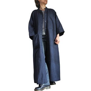 May include: A long, dark blue denim coat with a mandarin collar and button-up front. The coat has large pockets and three-quarter length sleeves. The coat is worn over a grey shirt and blue jeans.