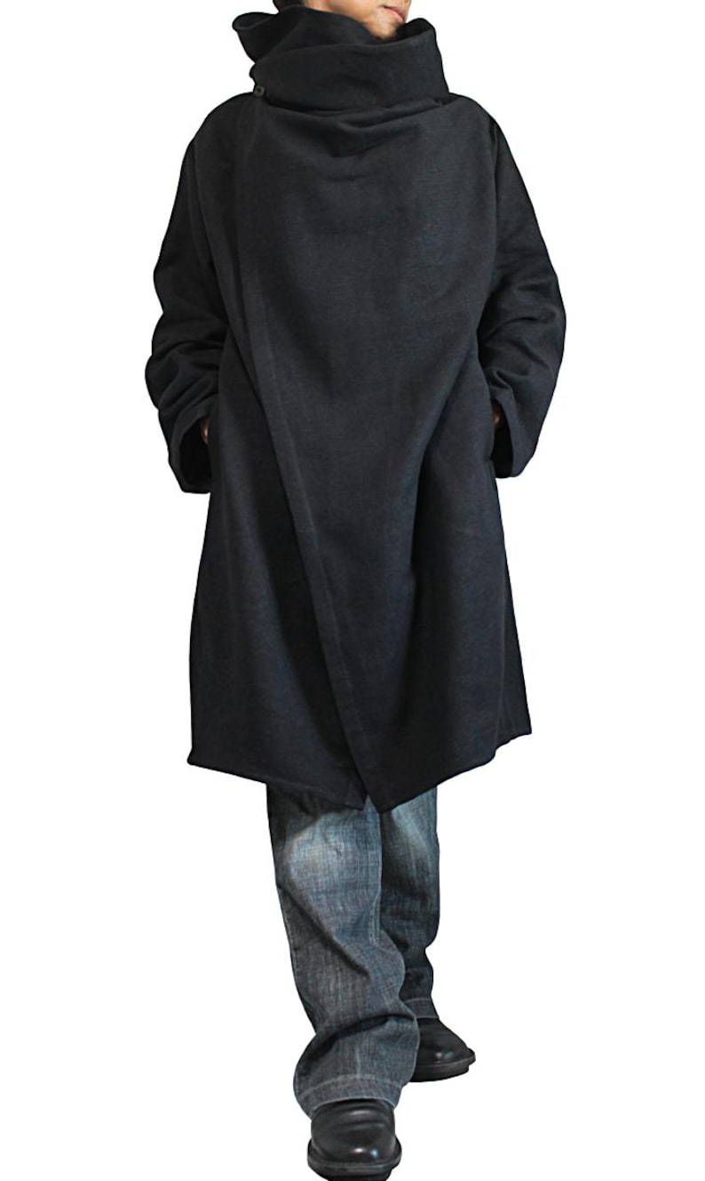 May include: A dark gray, long coat with a high, draped collar. The coat is knee-length and has a loose, flowing design. The person is wearing blue jeans and black shoes. The coat appears to be made of a soft, heavy fabric.
