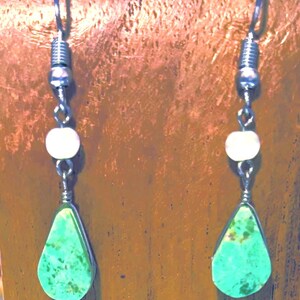 May include: A pair of dangle earrings featuring silver-toned metal hooks and wire components. Each earring includes a small, round, off-white bead positioned above a teardrop-shaped, bright green stone with dark brown mottled inclusions.