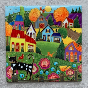 May include: A vibrant folk art painting showcases a village scene with houses of various colours. The artwork features houses with red, yellow, blue, and purple roofs, set against a green landscape with trees and flowers. A black cat with white spots is in the foreground.