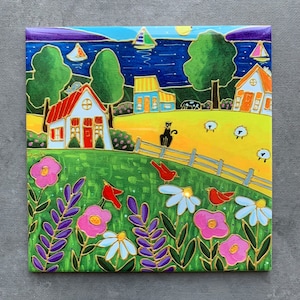 May include: A colourful ceramic tile featuring a whimsical illustration of a small town by a lake. The scene depicts houses with red roofs, a black cat, sheep, and a field of flowers. The tile has a bright yellow background and blue water.