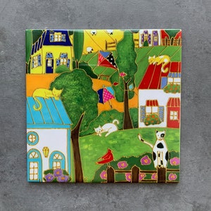 May include: A vibrant ceramic tile with a village scene. It shows houses with blue, yellow, and red roofs, green trees, and cats. A red bird sits on a fence, and a kite flies. The square tile is decorated with gold outlines and a playful design.