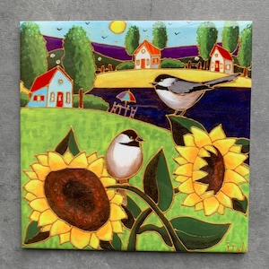 May include: Square ceramic tile featuring a vibrant landscape painting. The artwork depicts two chickadees perched on sunflowers, with a backdrop of houses, trees, and a lake under a sunny sky. The color palette includes green, yellow, blue, and red.