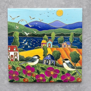 May include: Square ceramic tile featuring a colorful landscape painting. The artwork depicts a sunny day with birds, houses, and fields. Two birds perch on a flower-filled foreground. The sky is blue with a yellow sun.