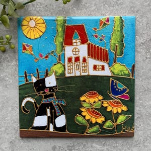 May include: A square decorative tile featuring a painted scene of a black and white cat wearing a blue and red scarf, sitting in a green field with yellow flowers, a butterfly, a white house with a red roof, a yellow sun, and kites.
