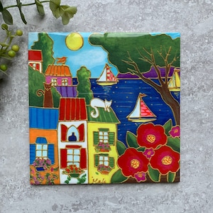 May include: A colorful illustration of a seaside town with houses, sailboats, and cats. The houses are painted in bright colors, including yellow, red, blue, and green. The sailboats are white with red and blue accents. The cats are black and white. The background is a blue sea with a green tree and a yellow sun.