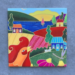 May include: A decorative ceramic tile depicting a landscape with a red bird, houses, fields, and a sailboat on a blue lake. The artwork uses bold colours and gold outlines, creating a vibrant piece for home decor.