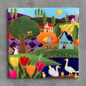May include: Square ceramic tile featuring a colorful, whimsical landscape scene. The artwork depicts a sunny day with a blue house, a lake with swans, and a field of tulips. A rabbit, birds, and a cat are also included in the design.