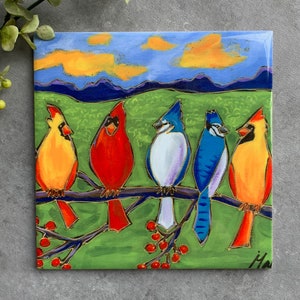 Peut inclure: Carreau de céramique carré avec une peinture de cinq oiseaux sur une branche. Deux cardinaux rouges et deux geais bleus sont sur une branche avec des baies rouges, sur un fond de champ vert, de ciel bleu et de nuages oranges.