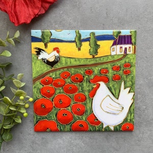 May include: A square ceramic tile featuring a painted landscape with a white chicken in the foreground and a black and white chicken in the background. The scene includes a field of red poppies, green grass, tall trees, a yellow meadow, and a small house.