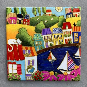 May include: Colorful square artwork depicting a coastal village scene. The painting features houses with various colored roofs and walls, a blue sea with sailboats, and a white cat resting on a red roof. The artwork has a whimsical style.