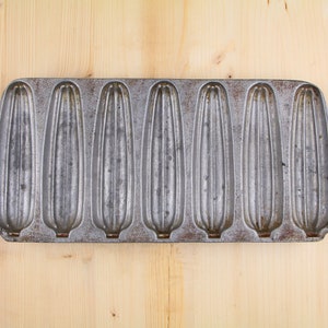 May include: A vintage metal cornbread pan with six individual molds. The pan is made of cast iron and has a rustic, aged look.