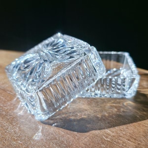 May include: A clear, square crystal trinket box with a lid, resting on a wooden surface. The box features intricate carved patterns and is partially open, revealing the interior. The sunlight creates shadows.
